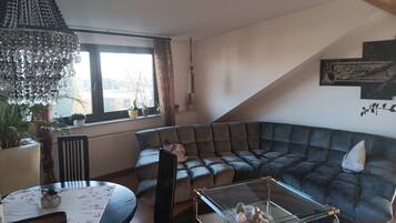 Een moderne woonkamer met een donkergrijze modulaire bank, een glazen salontafel, een eettafel met stoelen en een groot raam met gordijnen.