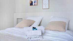 Una cama bien hecha con sábanas blancas y almohadas color beige, una toalla doblada y dos botellas de líquido azul sobre la cama.