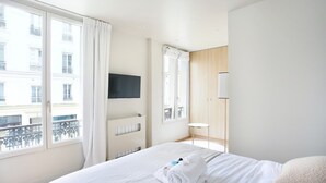 Habitación de hotel con cama, televisión montada en la pared, ventana con cortinas y vistas a edificios.