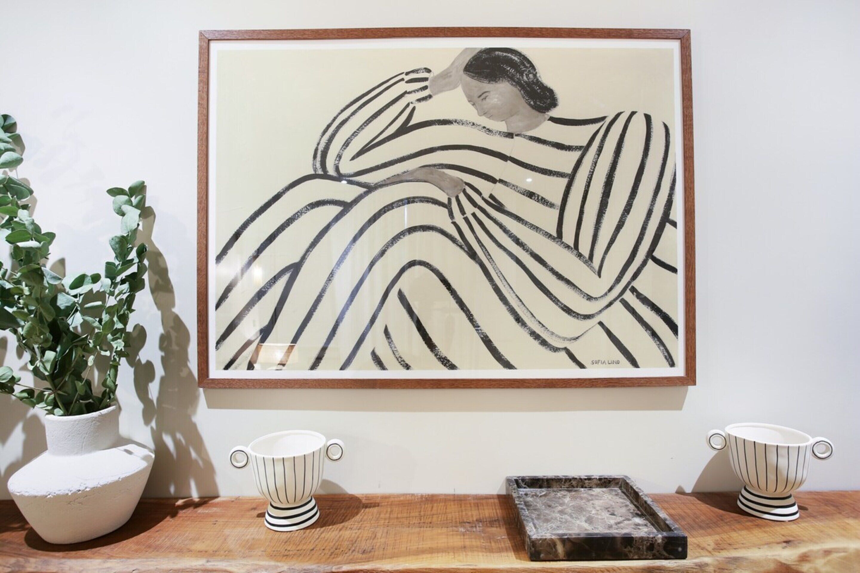 A framed artwork of a woman in a striped dress, a potted plant, and two striped cups on a wooden table.