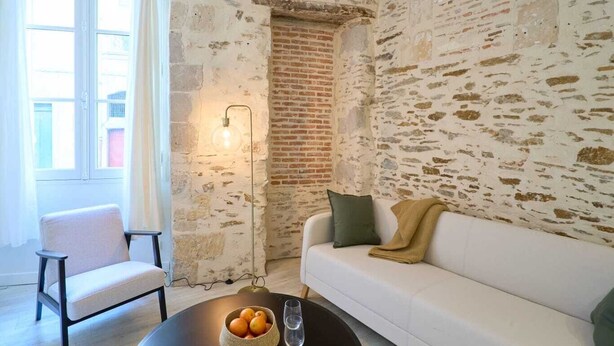 Una habitación con un sofá blanco, una silla, una mesa de centro con un frutero y vasos, una pared de ladrillo y una lámpara.