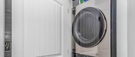 A laundry room with two stacked washers and dryers, a white door, and a shelf with laundry supplies.