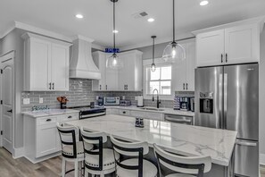 Cozinha moderna com armários brancos, uma ilha grande com banquetas, eletrodomésticos de aço inoxidável e iluminação pendente.