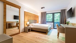 Una habitación de hotel moderna con suelo de madera, una cama grande, un escritorio con silla, televisor montado en la pared y un ventanal con cortinas.