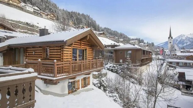 Una baita di legno con un balcone, immersa tra alberi e montagne innevate.