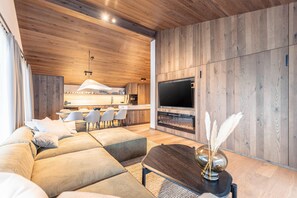 Uma sala de estar moderna com um amplo sofá modular, uma televisão de tela plana fixada numa parede de madeira, e um espaço de refeições com cadeiras, com uma cozinha ao fundo.