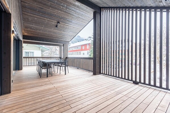Um deck de madeira com mesa e cadeiras, corrimão e vista para prédios e árvores ao fundo.