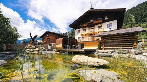 Ein traditionelles Holzchalet mit einem klaren Teich davor, umgeben von Felsen und Grünpflanzen.
