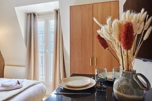 A neatly arranged hotel room with a bed, a dining table with plates and a vase of dried flowers, and a wardrobe in the background.