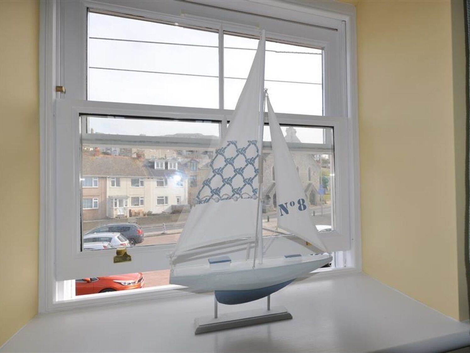 A model sailboat on a windowsill with a view of a street and buildings outside.