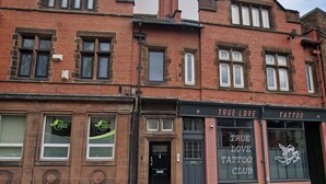 Ein rot backsteinsichtiges Gebäude mit einer schwarzen Tür und Fenstern. Rechts befindet sich ein Schild mit der Aufschrift 'True Love Tattoo Club'.