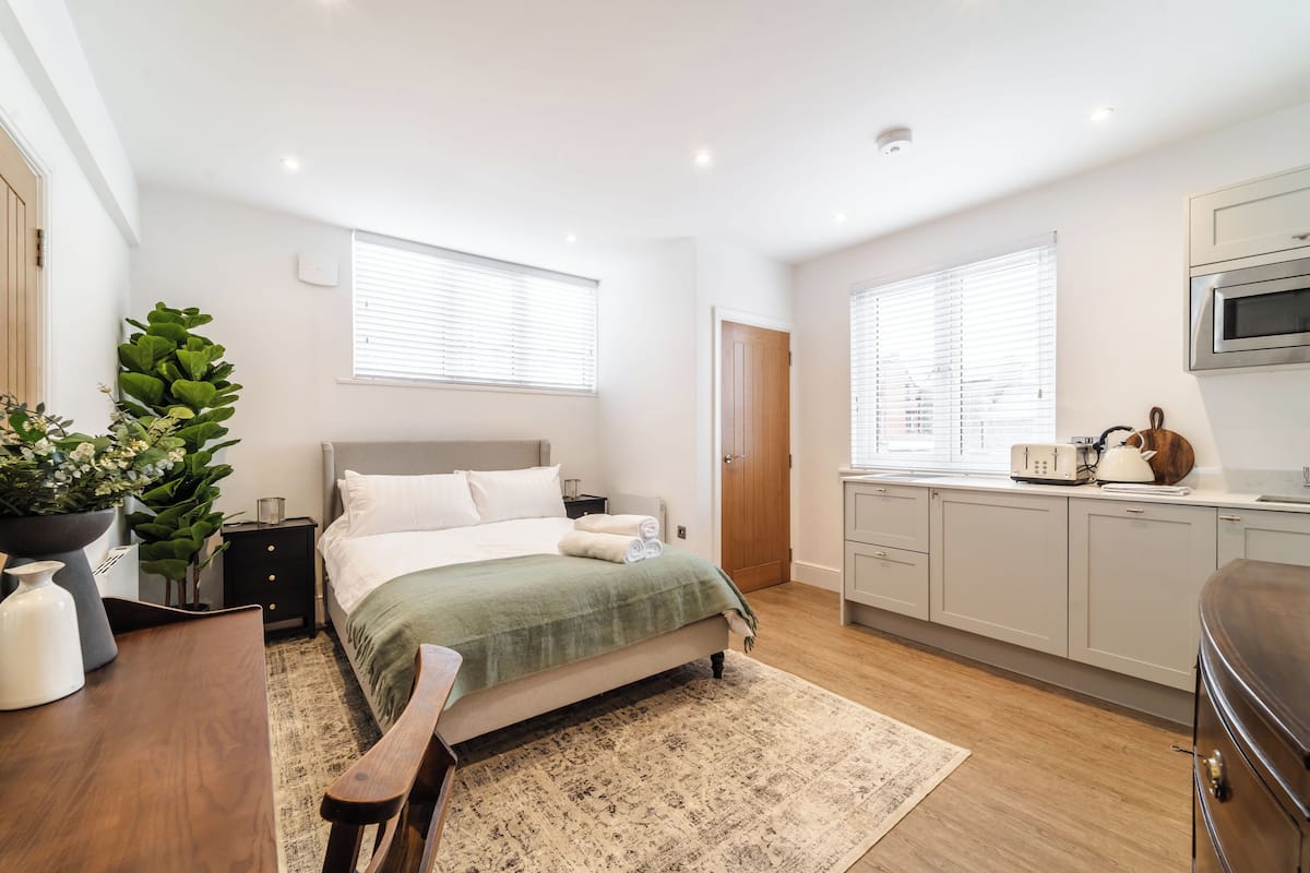 Une chambre moderne avec un lit, des tables de chevet, une petite table, une plante en pot et une vue sur la cuisine.