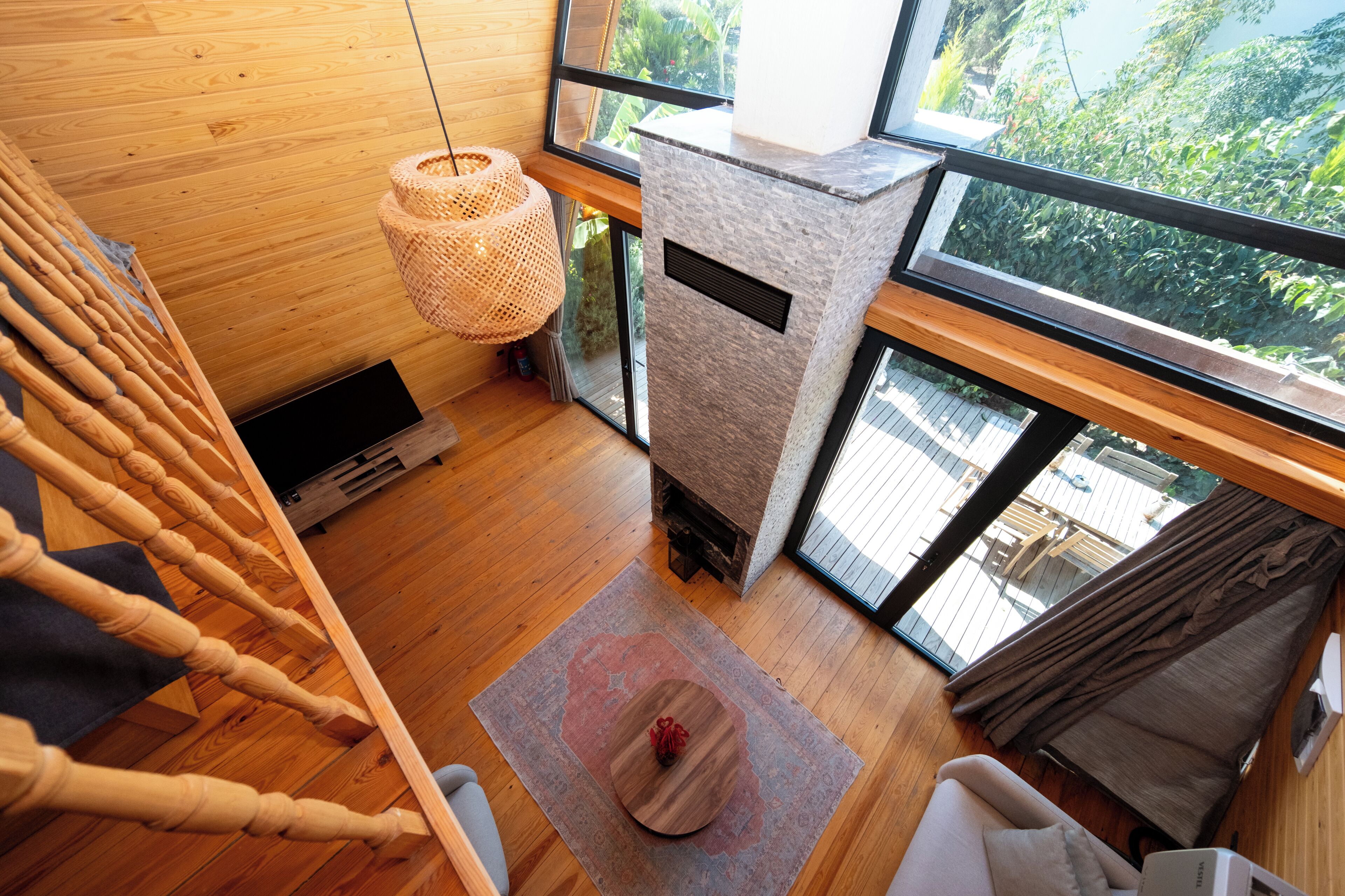 A room with a wooden floor, a stone fireplace, a large window with a view of greenery, a woven light fixture, and a small rug.
