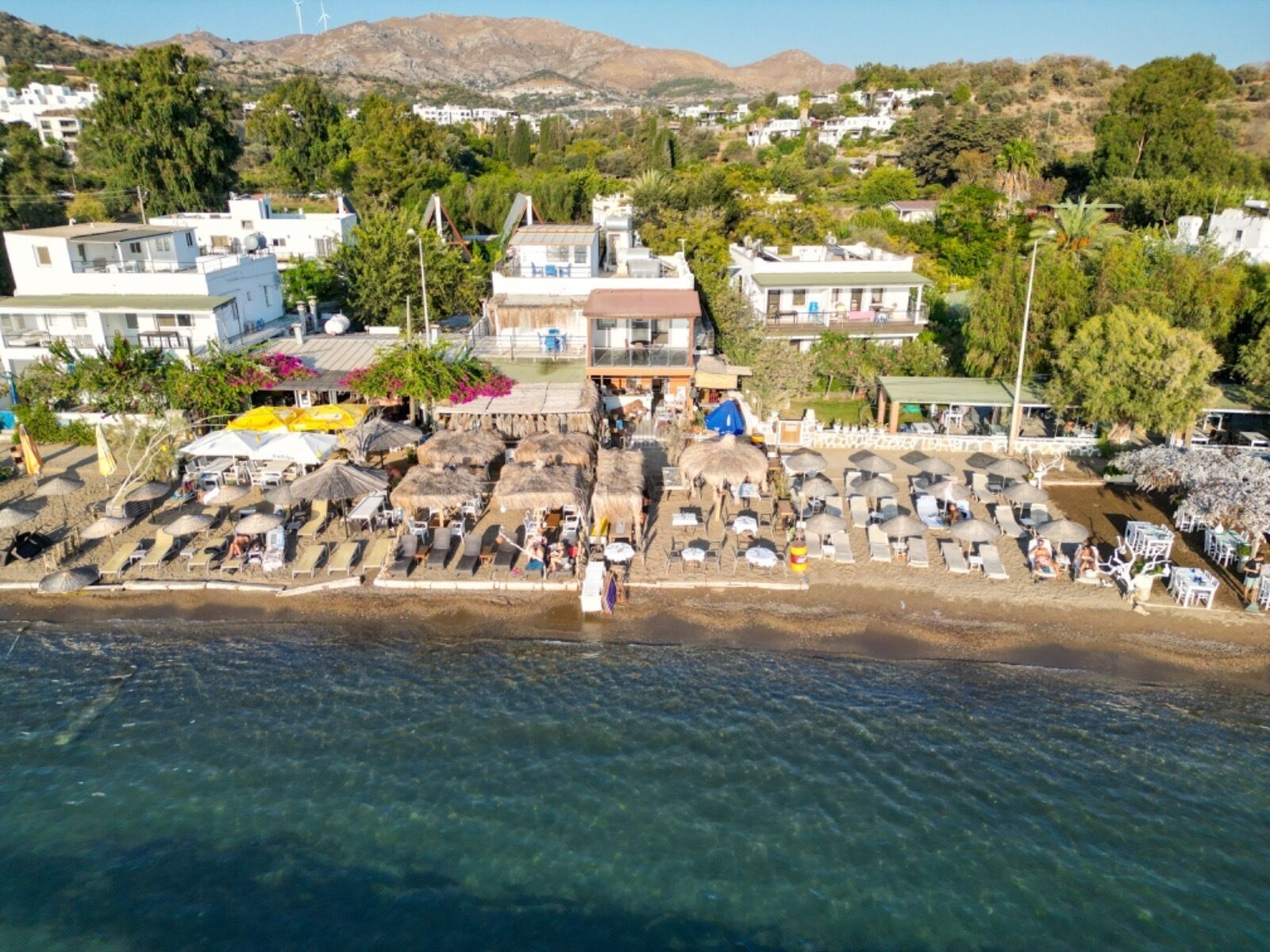 An aerial view of a coastal resort with beach umbrellas, sun loungers, and a building with a balcony overlooking the beach.