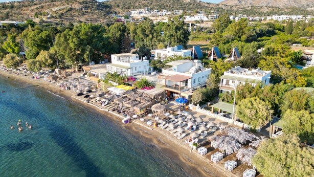 An aerial view of a coastal area with buildings, a beach, and a clear blue sea. - BEGONVİL GÜMÜŞLÜK  (Bodrum)