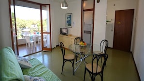 A living room with a glass dining table, black chairs, a green sofa, and a balcony with white chairs. - Fantastic Flat With Pool View Just Steps From the sea (Lignano Sabbiadoro)