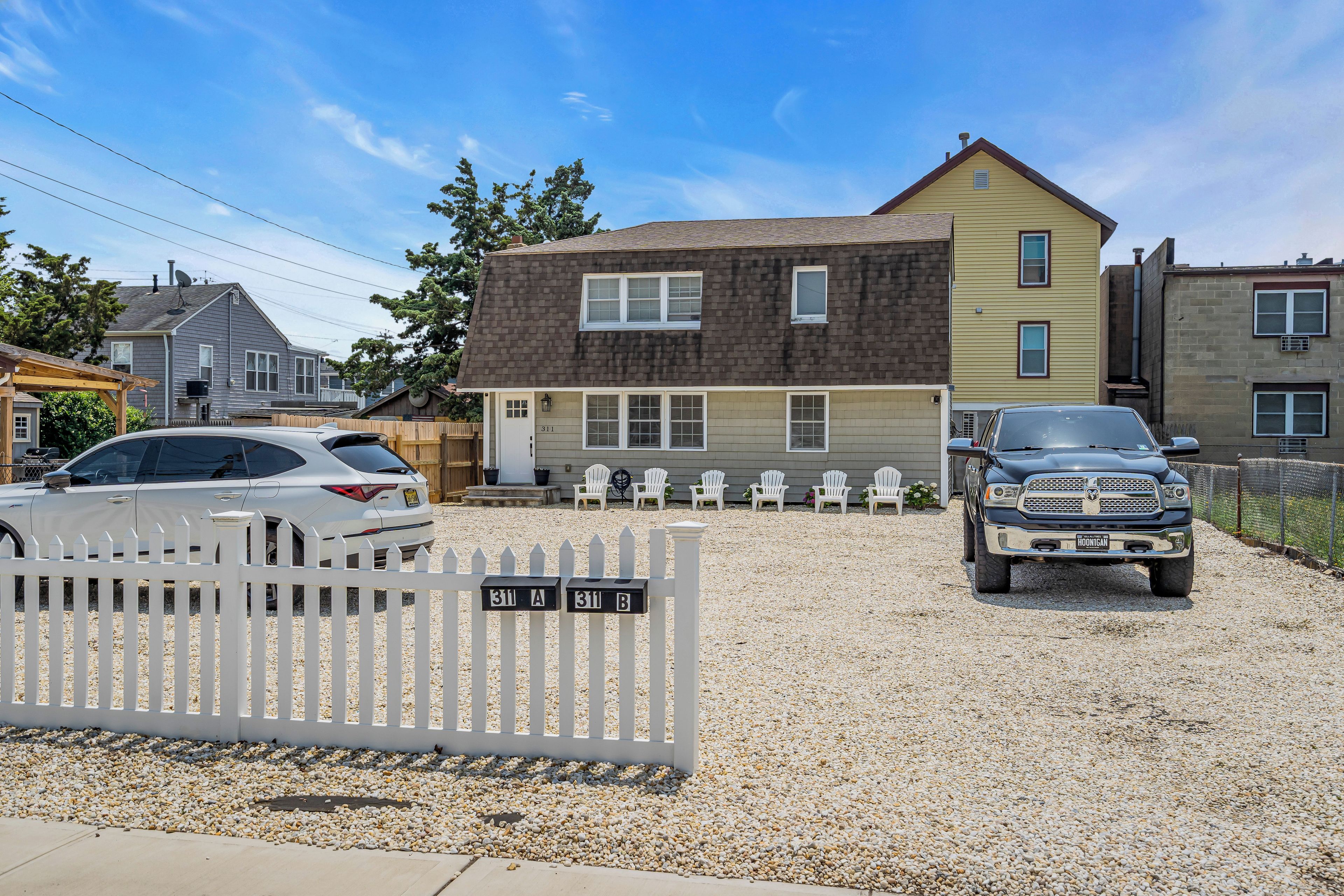 A residential driveway with a gravel surface, a white picket fence, and two parked cars.