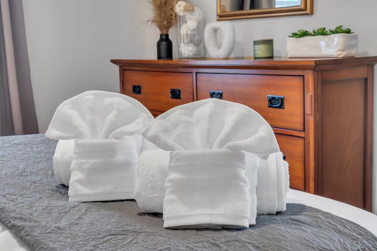 A neatly made bed with two white towels folded on top, a wooden dresser with drawers, and a vase with dried plants on top.