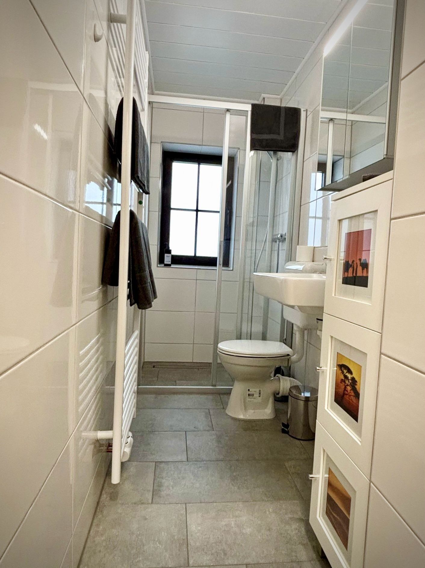 A compact bathroom with a toilet, a sink, and a shower. There is a window providing natural light, and the walls are tiled.