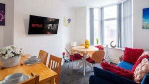 Een moderne woonkamer met een eethoek, een flatscreen televisie aan de muur en een groot raam met gordijnen.