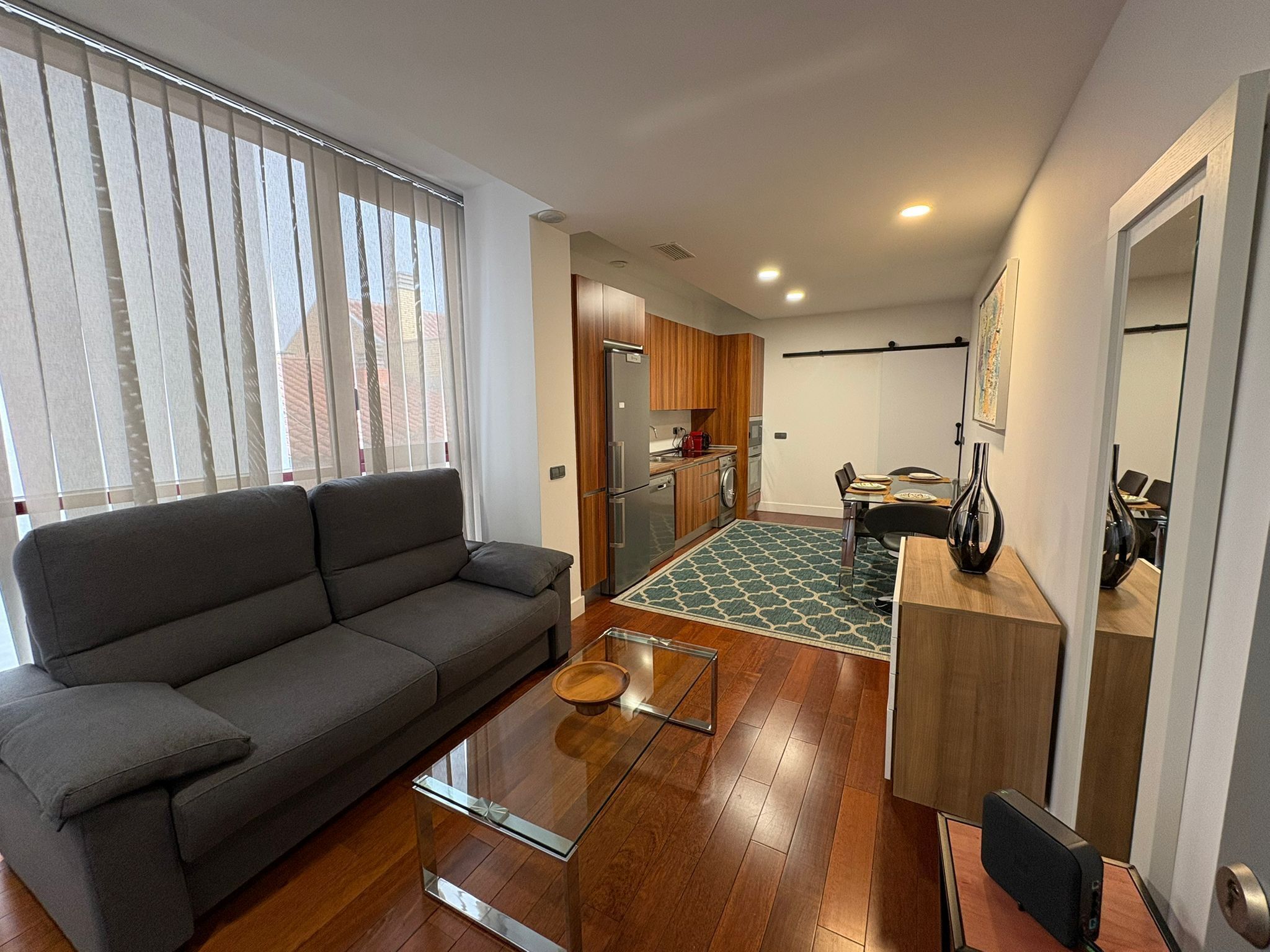 Un apartamento moderno con un sofá gris, una mesa de centro de vidrio, una cocina con armarios de madera y un comedor con mesa y sillas.