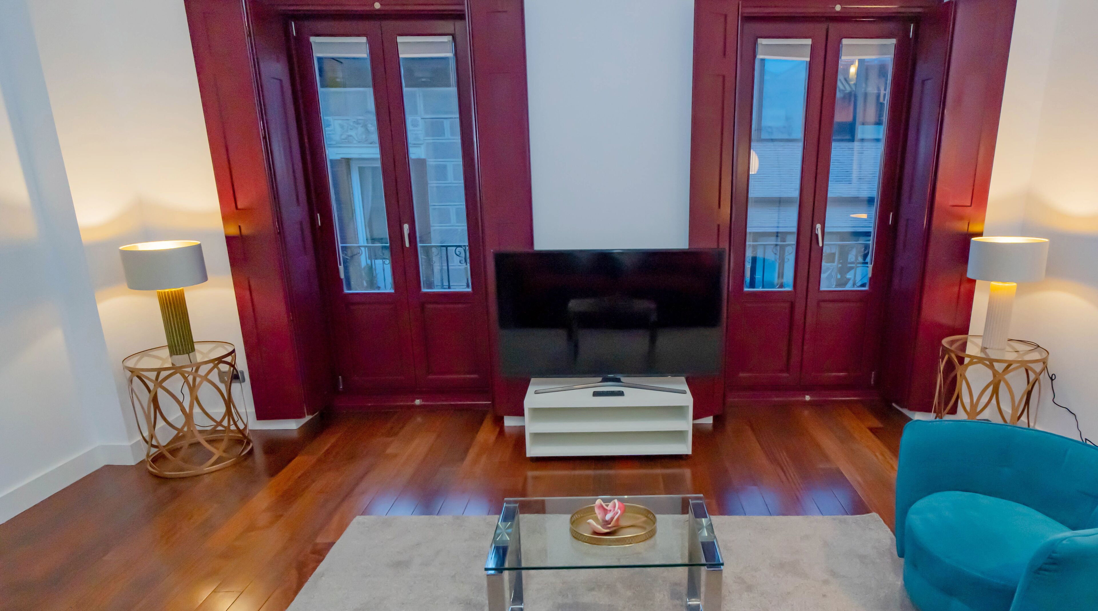 Un salon doté d’un téléviseur à écran plat, d’un parquet, de portes rouges et de deux lampes posées sur des tables d’appoint.