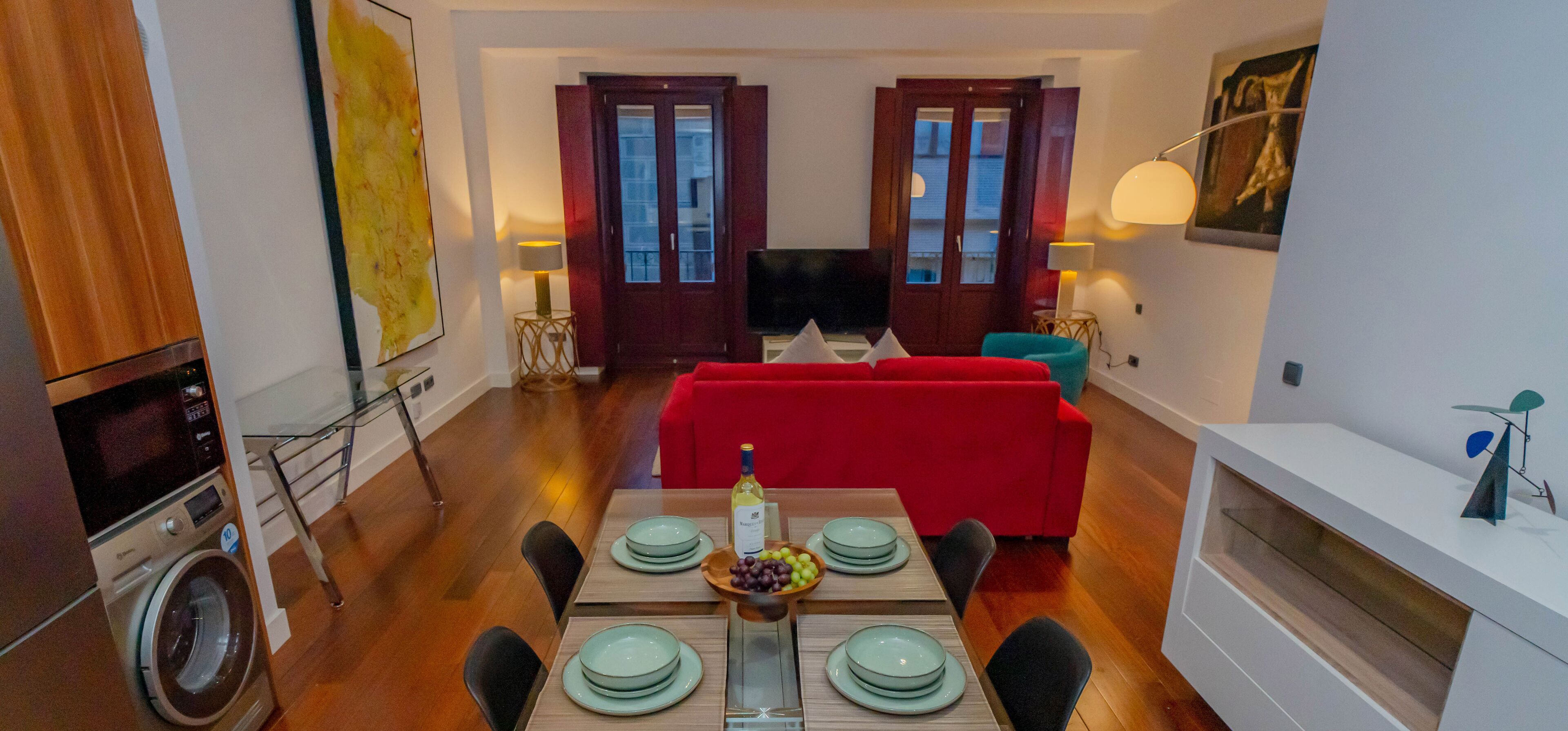 A modern living room with a red sofa, a dining table set with plates and a bottle, and a TV in the background.