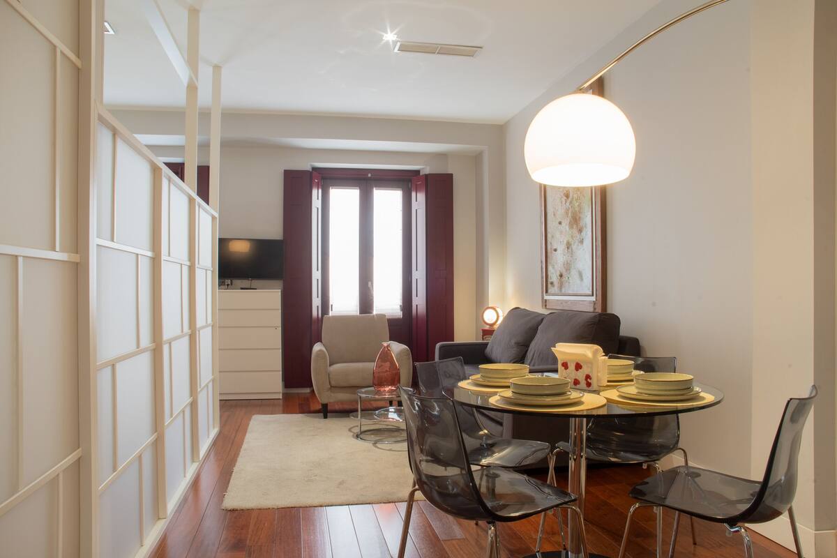 A modern living room with a dining area, featuring a sofa, chairs, a table set with plates, and a television in the background.