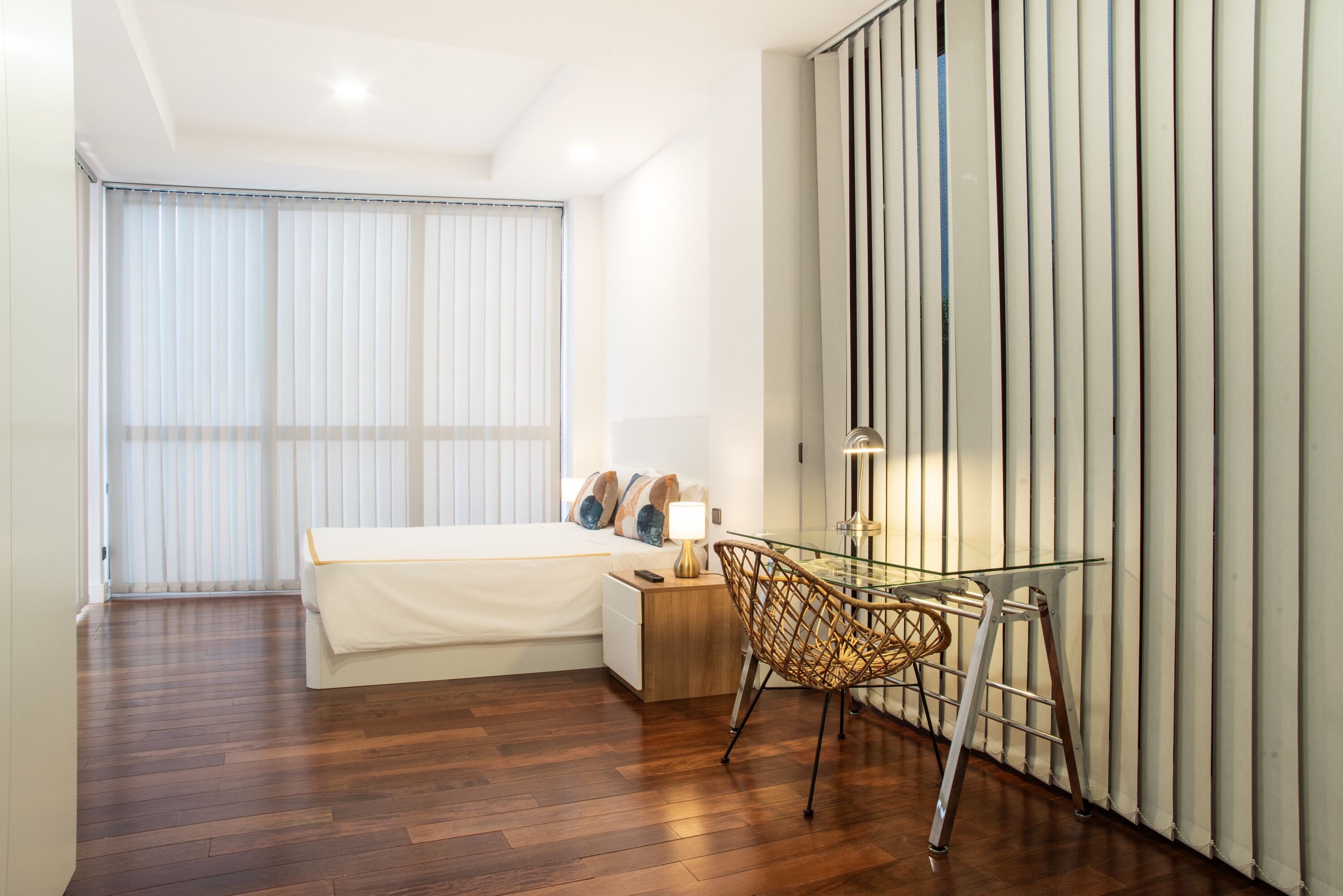Un dormitorio moderno con suelo de madera, una cama con ropa de cama blanca, un escritorio de vidrio con una silla y un ventanal con persianas.