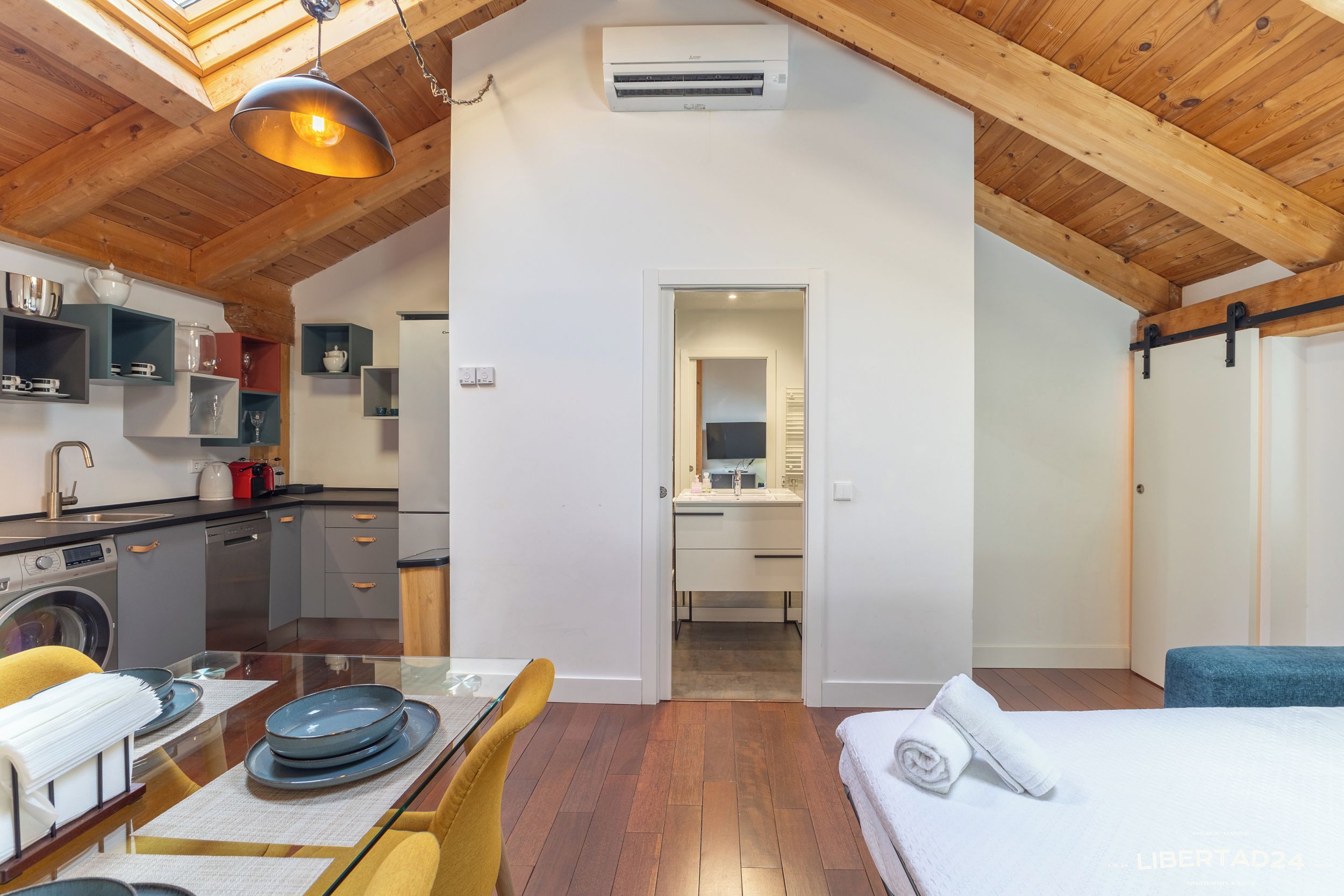 A compact kitchen with modern appliances, a dining area with a table set for two, and a bedroom with a bed and a blue chair.