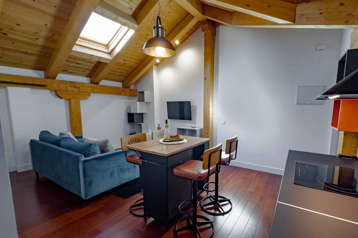 A cozy loft-style room with a wooden ceiling, a blue sofa, a dining table with bar stools, and a kitchen area with a stove.