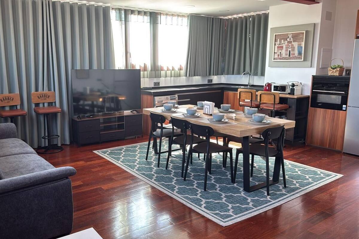 A modern living room with a sofa, TV, dining table, chairs, kitchen area, and a rug.