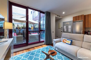 A modern living room with a sofa, a coffee table, and a large sliding glass door leading to an outdoor area with plants and a table. - Libertad 24 (Madrid)