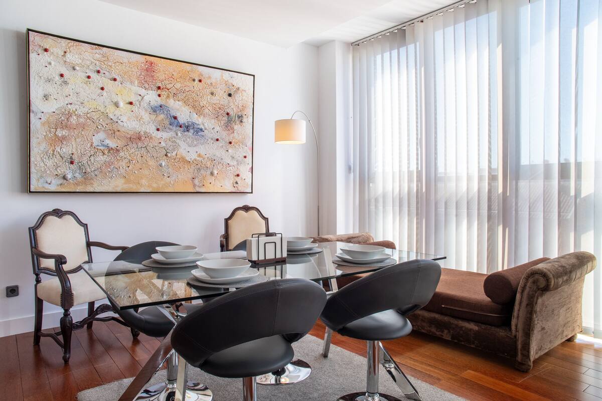 A modern living room with a glass dining table, black chairs, a brown sofa, and a large abstract painting on the wall.