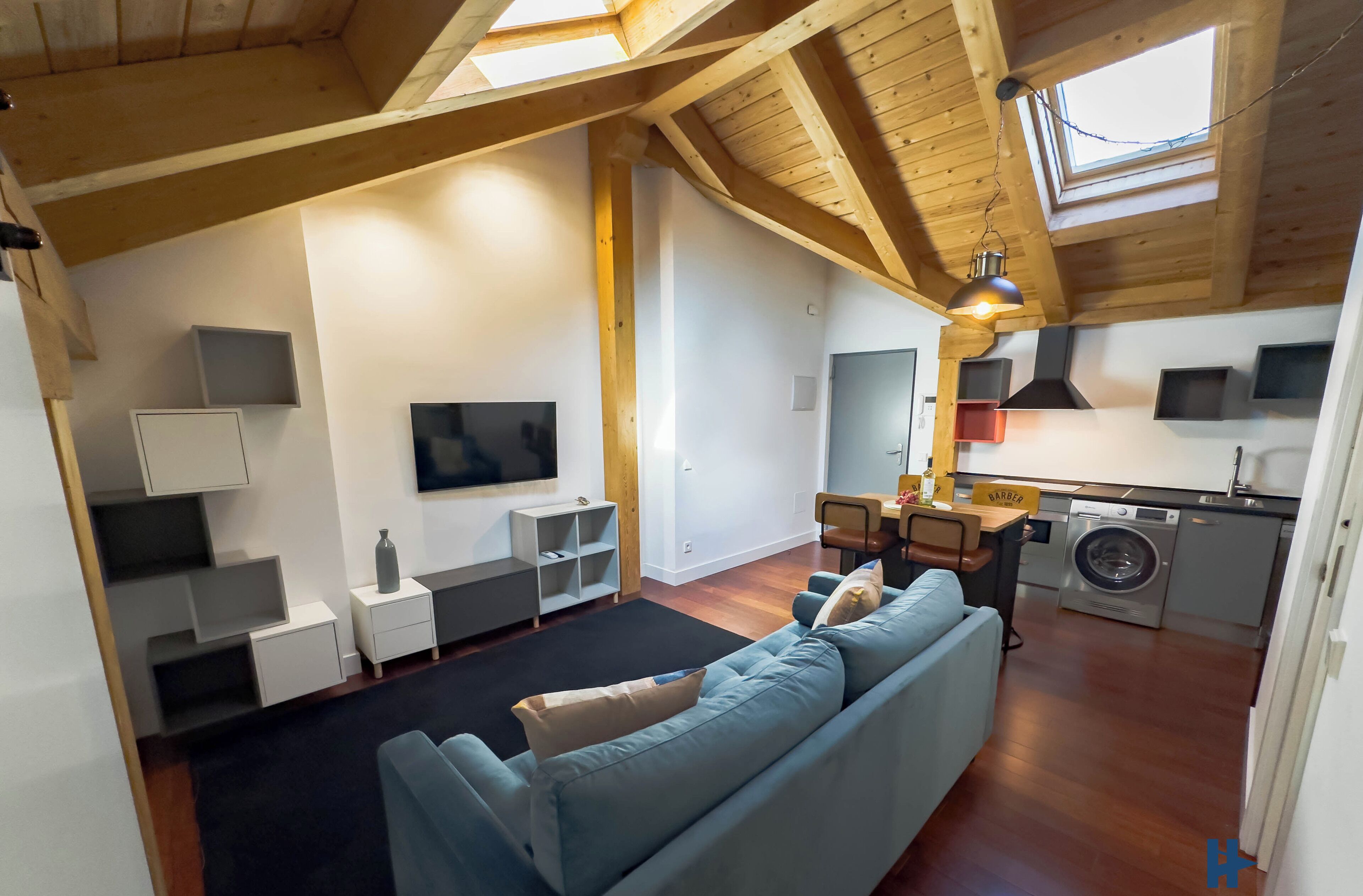 A modern living room with a sofa, TV, and built-in shelves. A kitchen area with a washing machine is visible in the background.