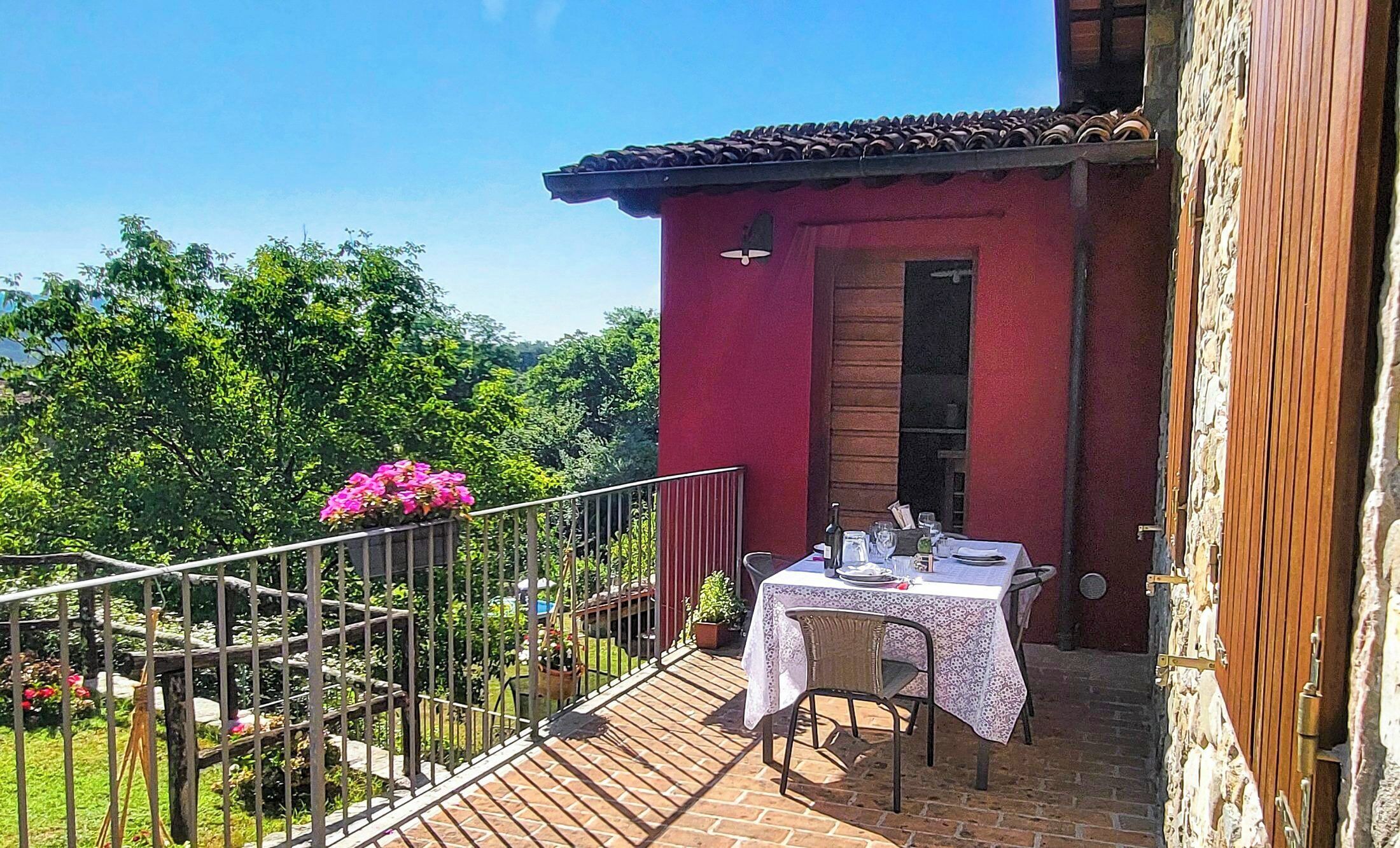 Un edificio rojo con una mesa en el balcón dispuesta para comer, rodeado de follaje y un cielo despejado.