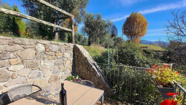 Un mur de pierre, une table en bois avec une bouteille, des verres et une assiette de nourriture, et une rambarde en métal ornée de plantes.