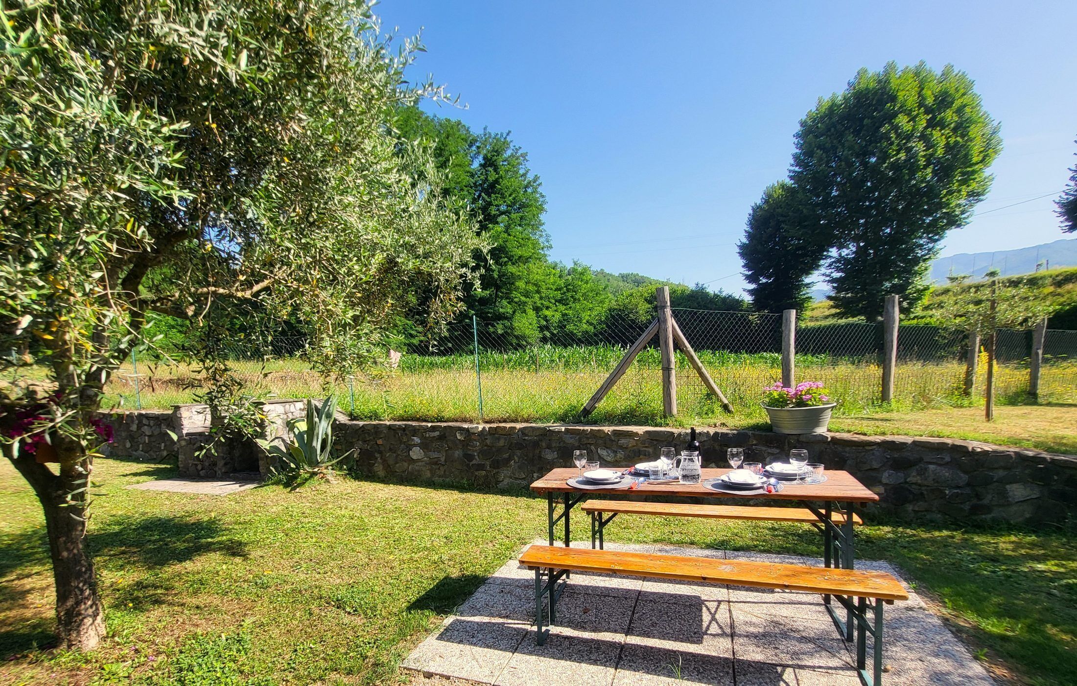 An outdoor dining area with a stone wall, a wooden table set with plates, glasses, and a flower pot.