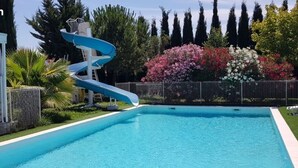 A swimming pool with a blue slide, surrounded by a fence, palm trees, and flowering bushes. - Gîte Tropica Nîmes air-conditioned Swimming pool, slide, straw hut (Nîmes,Gard,)