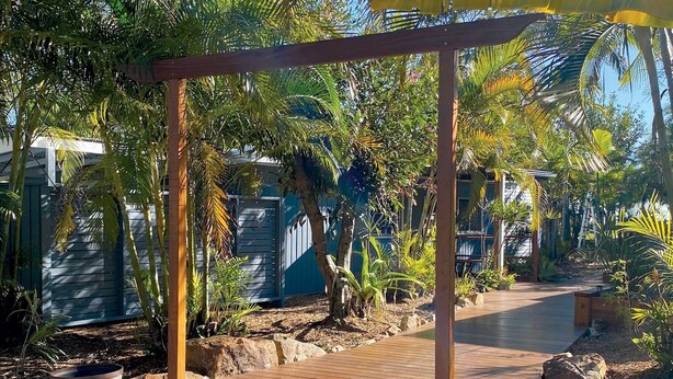 Une structure de pergola en bois, entourée de plantes tropicales, et un chemin en bois.