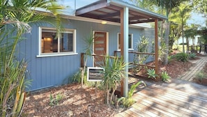 Une petite maison de plain-pied avec un porche couvert, entourée de plantes tropicales et dotée d’une terrasse en bois.