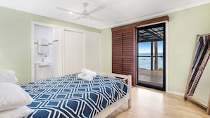 A bedroom with a bed, a ceiling fan, a bathroom with a toilet and sink, and a door leading to a balcony with a view of the ocean.