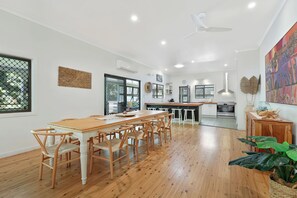 A spacious dining area with a wooden table and chairs, a kitchen in the background, and a large window with a view of greenery.