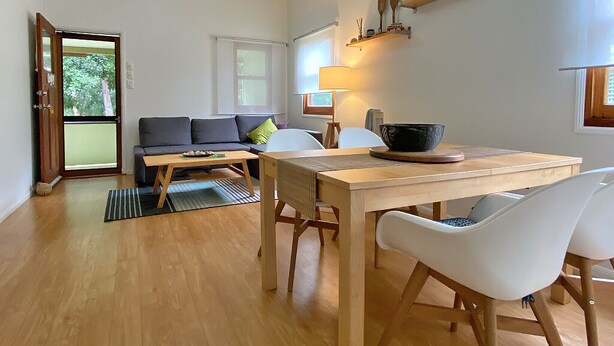 A modern living room with a sofa, a dining table, and chairs. There is a lamp on the table and a window with a view of trees.