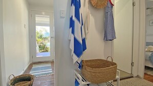 A hallway with a wooden floor, a white door, and a view of the outdoors. There are striped towels and a straw hat hanging on the wall.