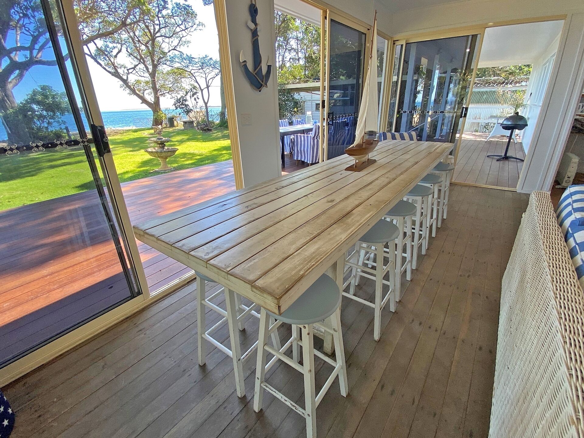 Una mesa de comedor de madera con taburetes blancos, ventanas amplias con vistas al océano y un sofá de mimbre.