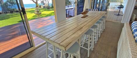 Una mesa de comedor de madera con taburetes blancos, ventanas amplias con vistas al océano y un sofá de mimbre.