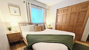 A neatly arranged bedroom with a bed, bedside tables, a wooden wardrobe, a window with curtains, and a framed picture on the wall.