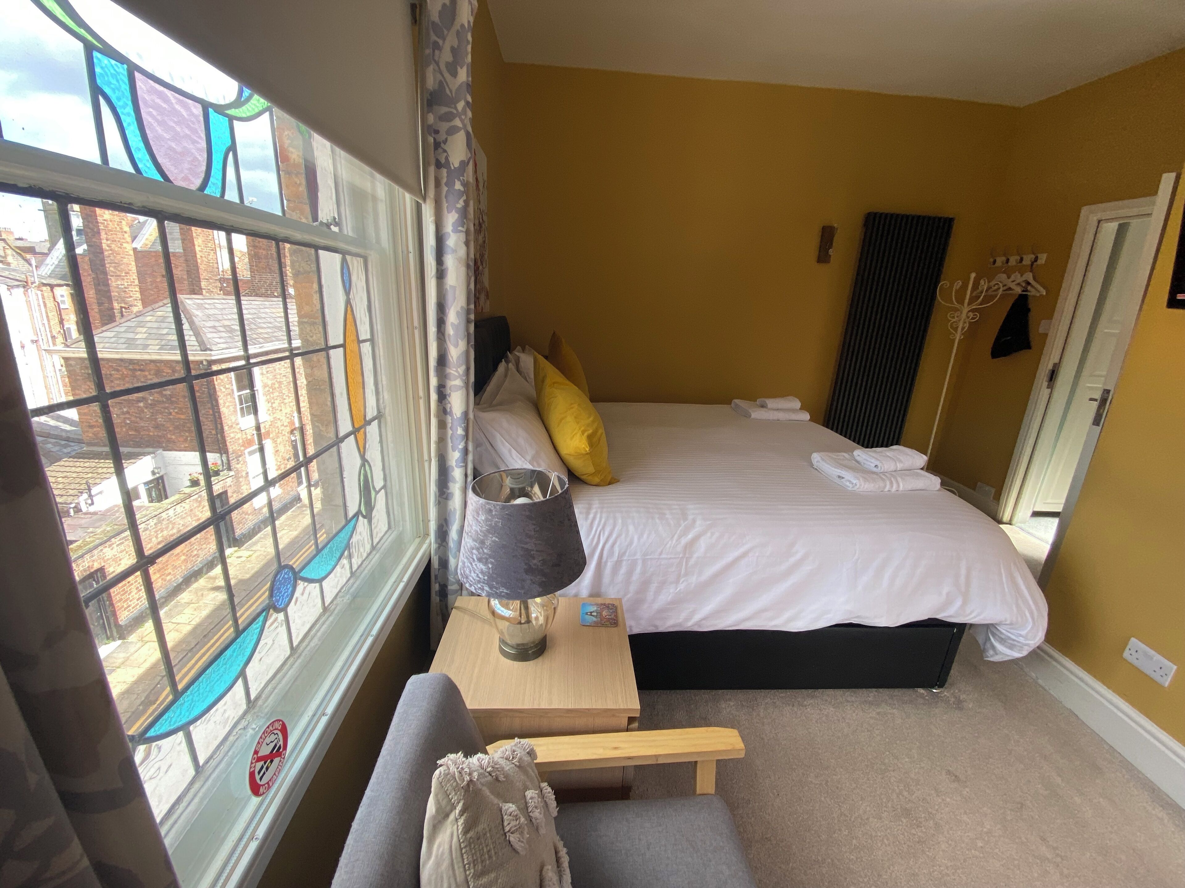 A hotel room with a bed, a bedside table with a lamp, a window with a view of buildings, and a door.