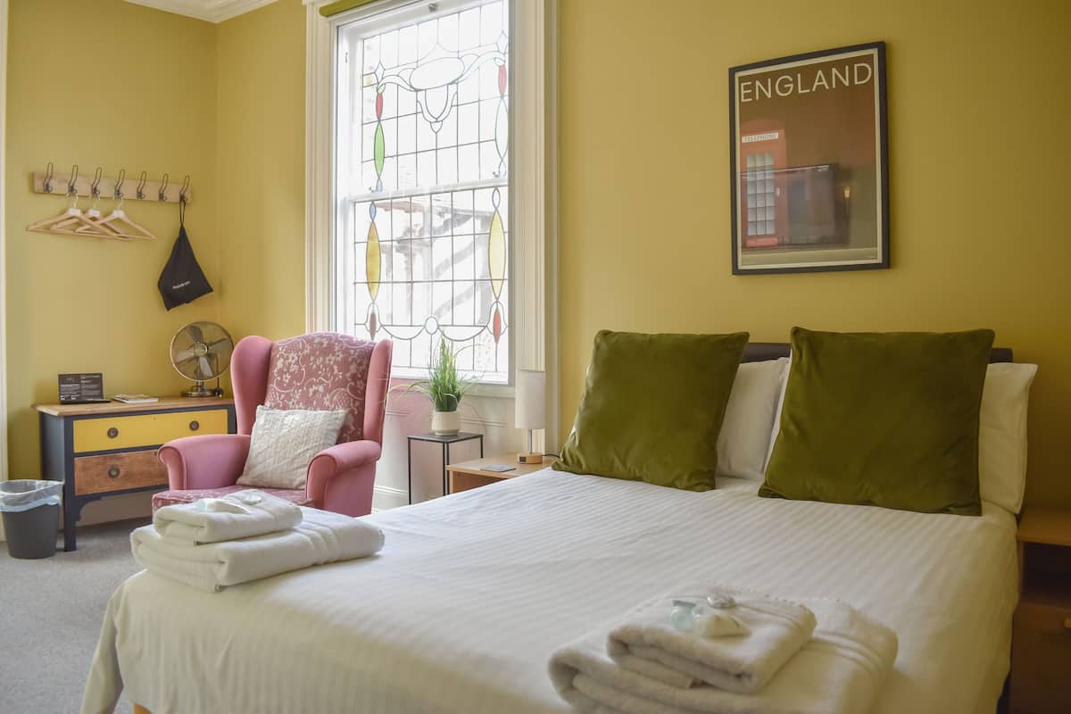 A hotel room with a bed, a chair, a bedside table with a lamp, a fan, and a window with a view of England.